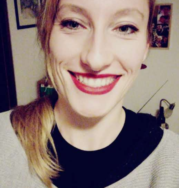 Lächelnde Frau mit roten Lippen und blonden Haaren, in einem Wohnraum.