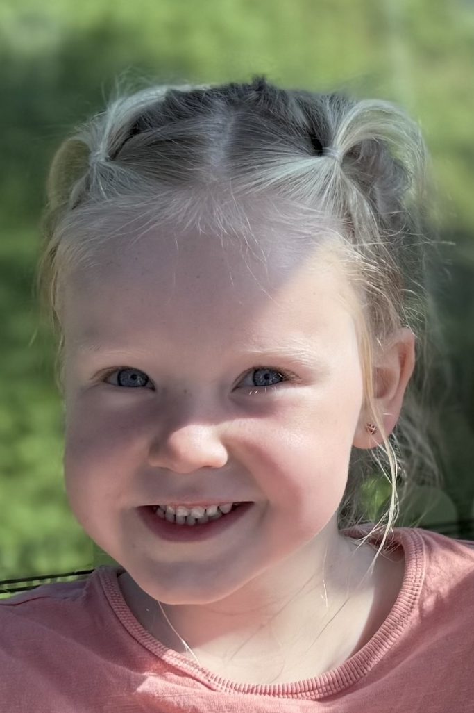 Lächelndes kleines Mädchen mit blonden Haaren und einem rosa T-Shirt.