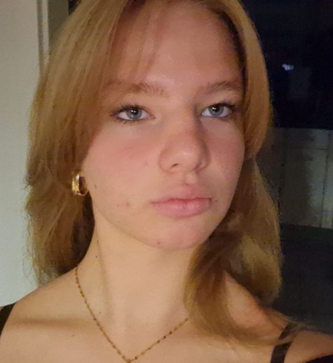 Junge Frau mit blonden Haaren und blauen Augen, serösem Gesichtsausdruck.